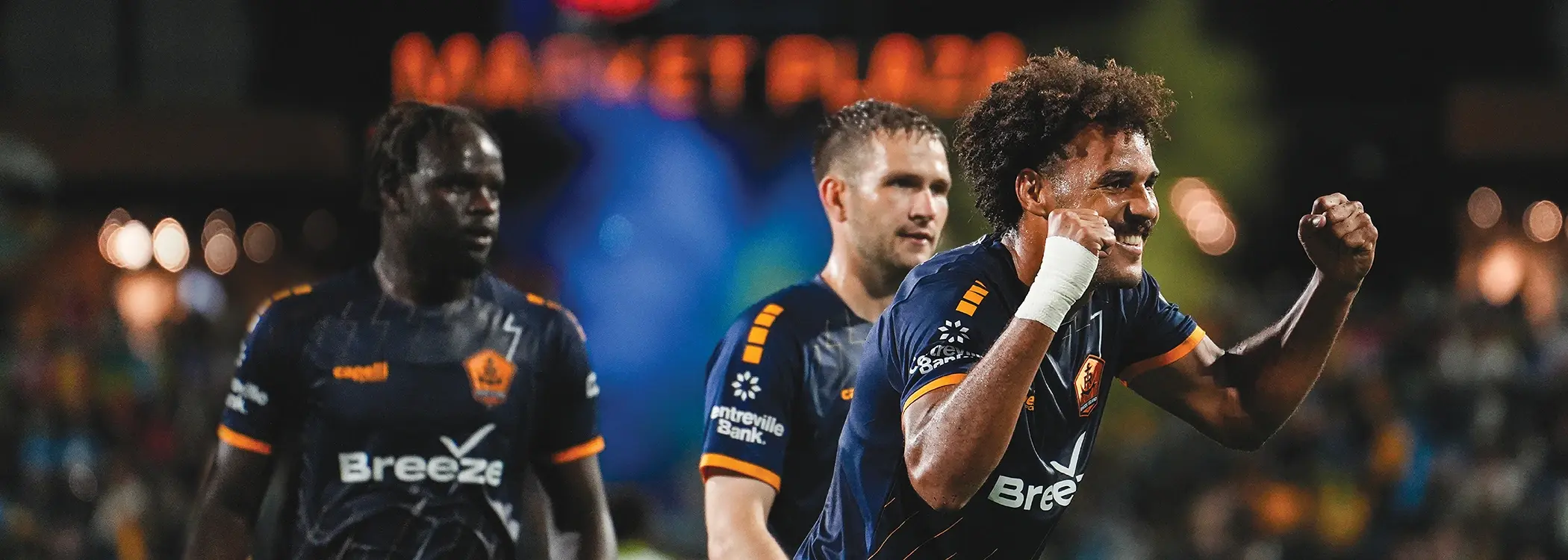 Three professional rugby players in dark jerseys with sponsor logos celebrating during a night match, showing emotion and team spirit on the field.