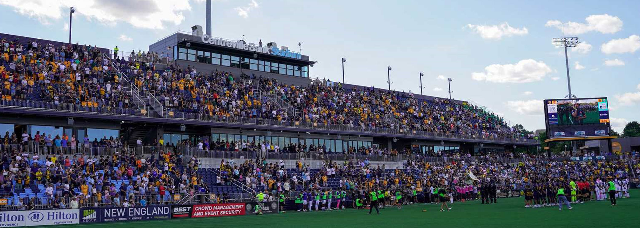 Crowded stadium grandstand and players lined up along the field edge.