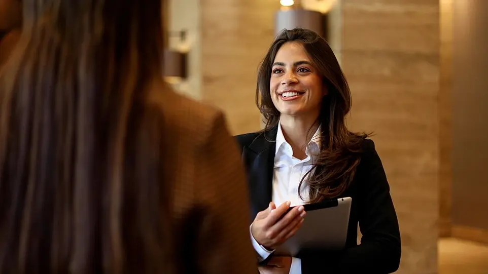 A smiling businesswoman in a blazer holds a tablet while talking with a colleague in an office lobby.