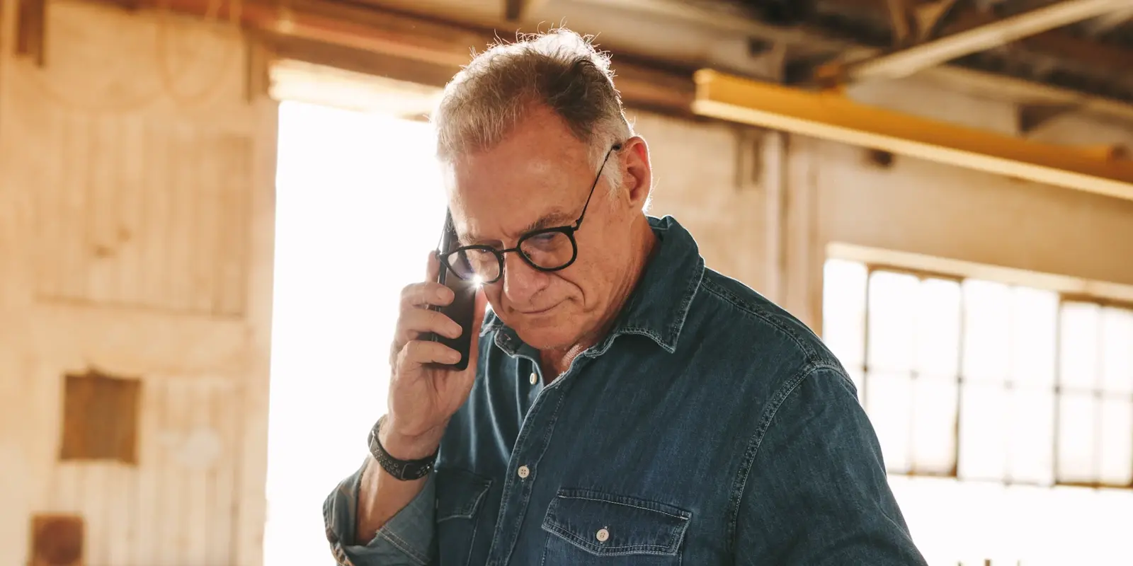 Mature man with glasses wearing denim shirt talking on phone while working on laptop at workbench in workshop