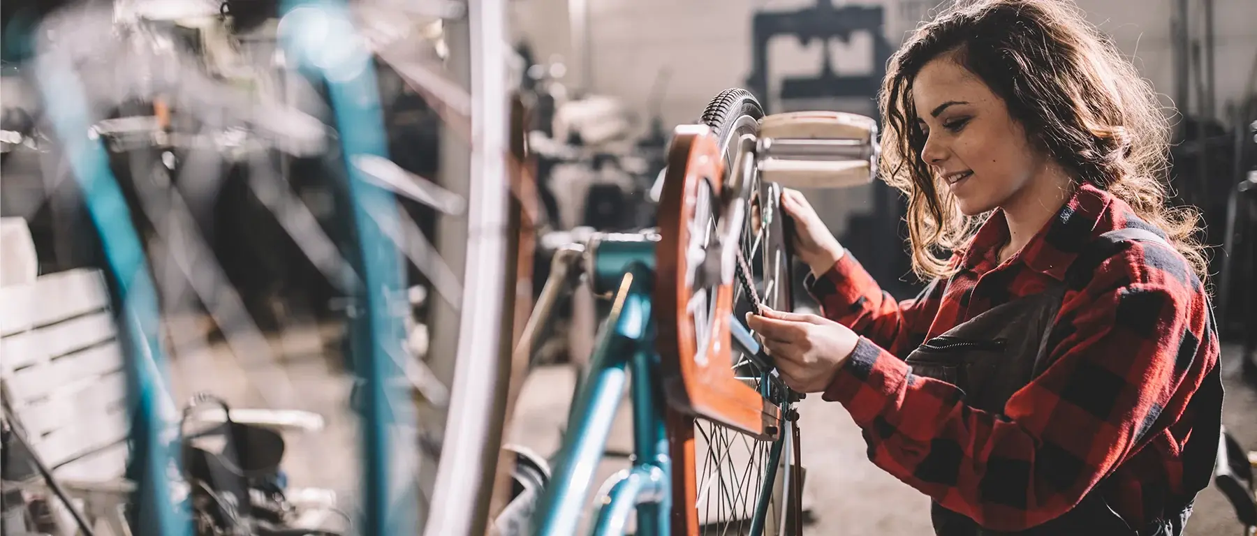 A woman in a red plaid shirt adjusts the handlebars of a bicycle in a repair shop, with more bikes racked around her.