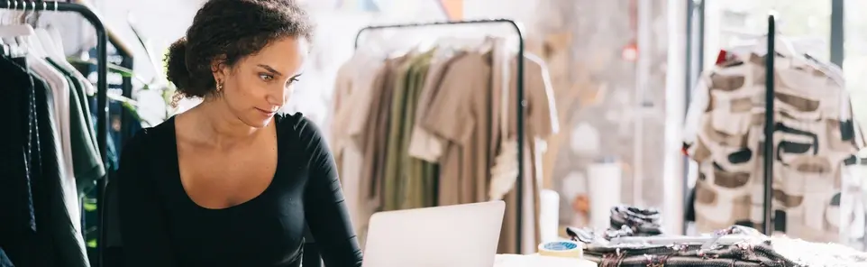 A boutique owner reviews her laptop at the checkout counter, surrounded by clothing racks in her shop.