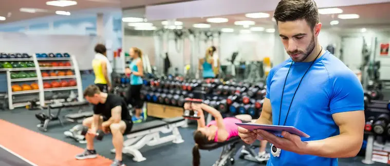 A personal trainer reviews a tablet in a gym while clients exercise with dumbbells in the background.