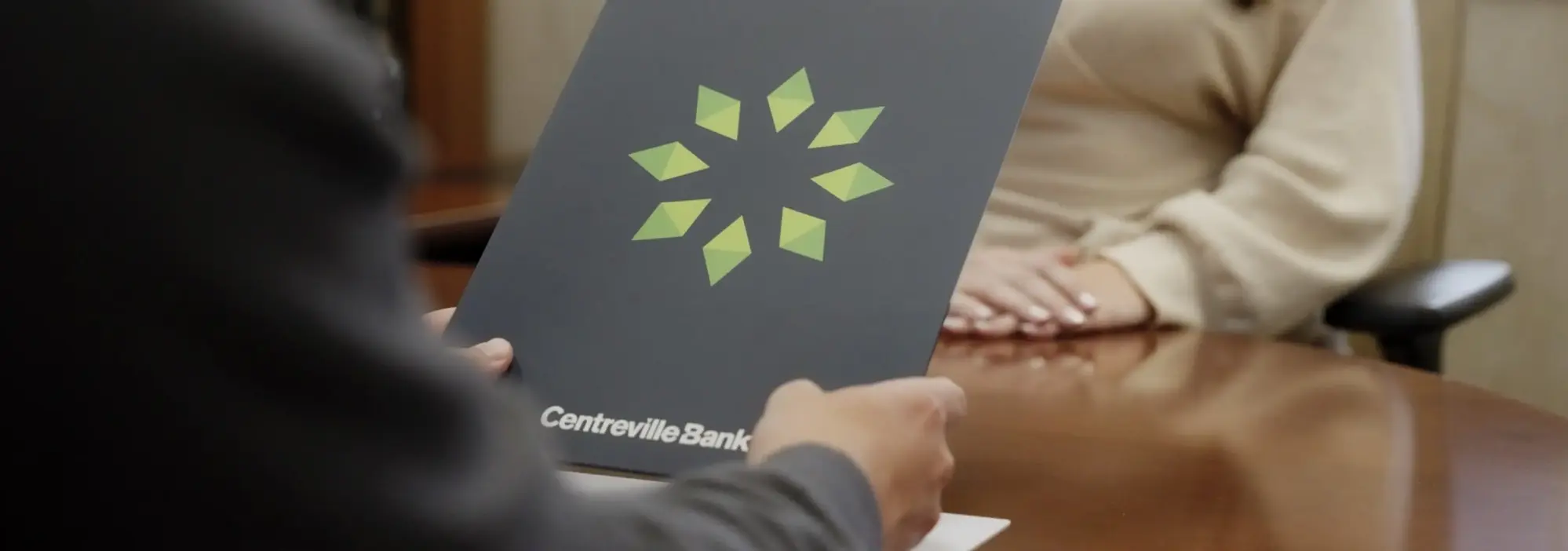 Two professionals in an office setting exchanging a folder with a green leaf logo design, representing sustainability or environmental initiatives