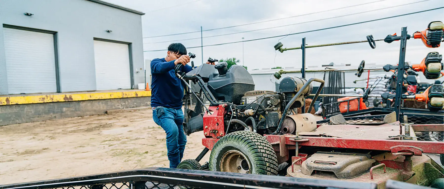 A landscaping business owner uses his phone beside a trailer loaded with lawn equipment outside his shop.