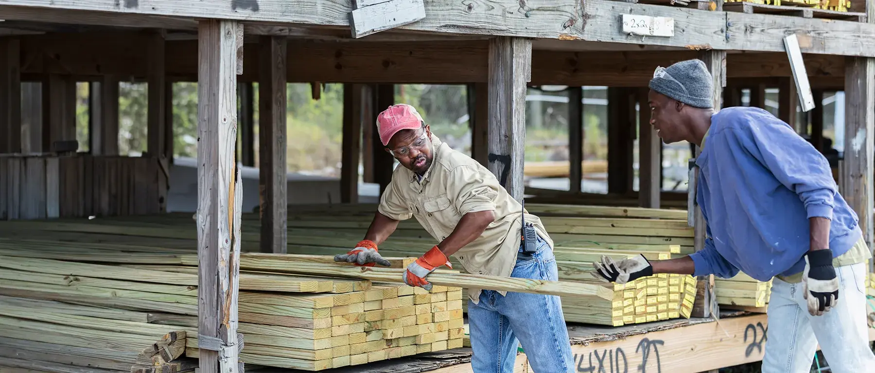 Two men in work clothes lift a bundle of fresh lumber together at a building site, under the timber frame of a raised structure.