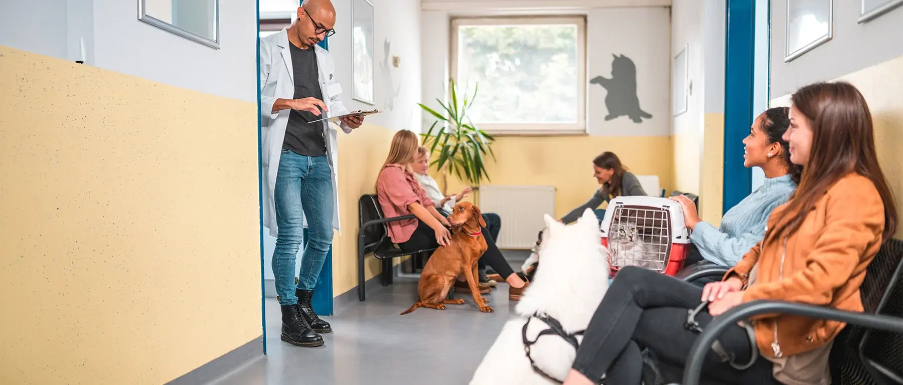 A veterinarian holding a clipboard stands in a clinic waiting room where seated clients wait with two dogs and a pet carrier.
