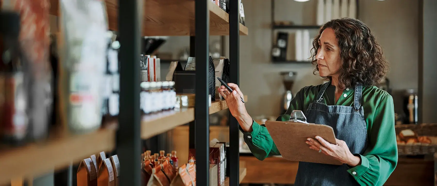 A woman in a green apron checks stock against a clipboard in a shop, reaching toward bottles and jars on tall wooden shelves.