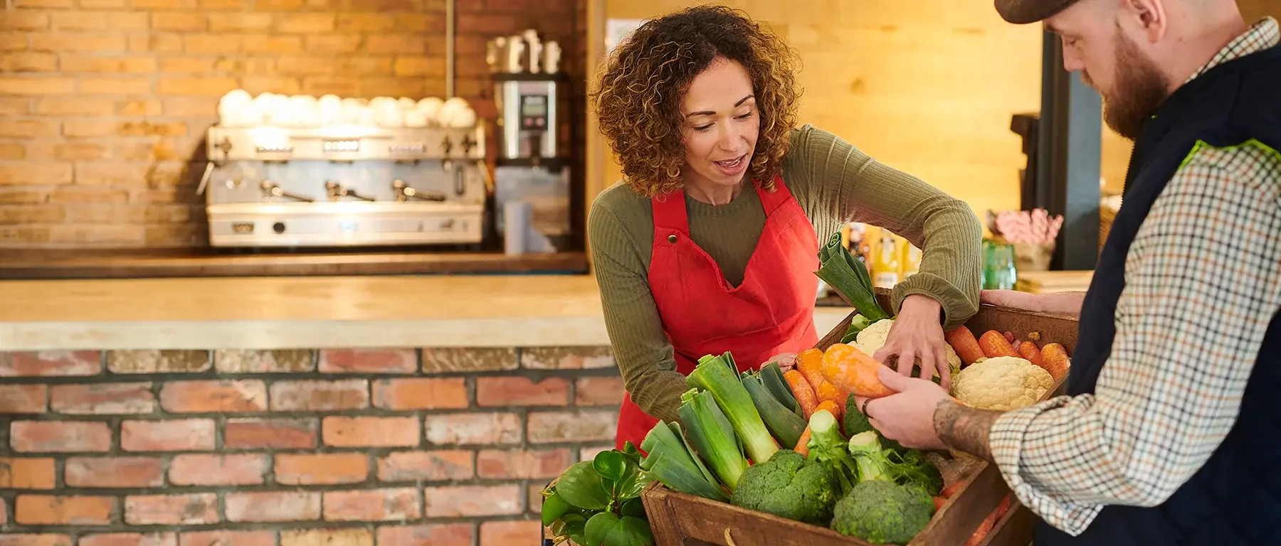 A market vendor in a red apron shows fresh produce to a customer at a wooden stall, with an espresso bar and exposed brick behind them.
