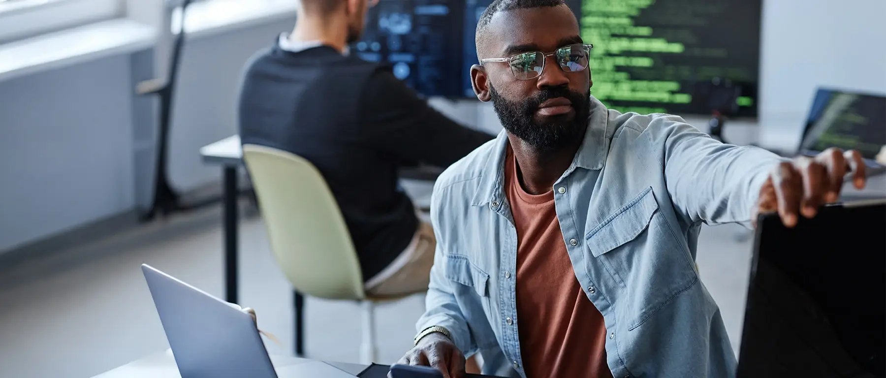 A man in glasses and a denim shirt works at a laptop in an open office, with a colleague and a monitor of code behind him.