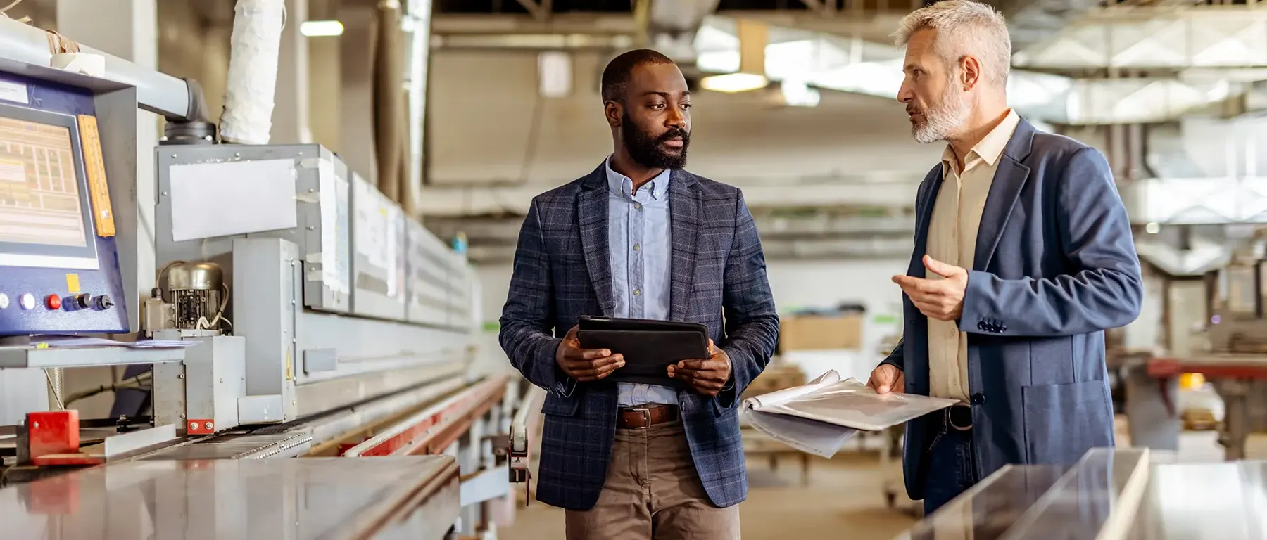 Two business professionals in blazers review a tablet and documents while walking through a manufacturing facility.
