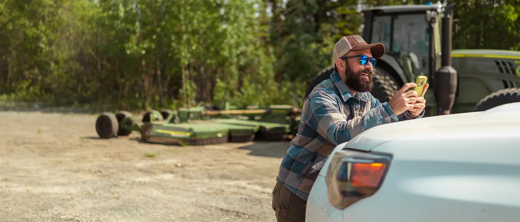 A man in a baseball cap, sunglasses and a plaid shirt leans against the back of a white pickup truck looking at his phone, with green farm machinery and trees behind him in a gravel yard.