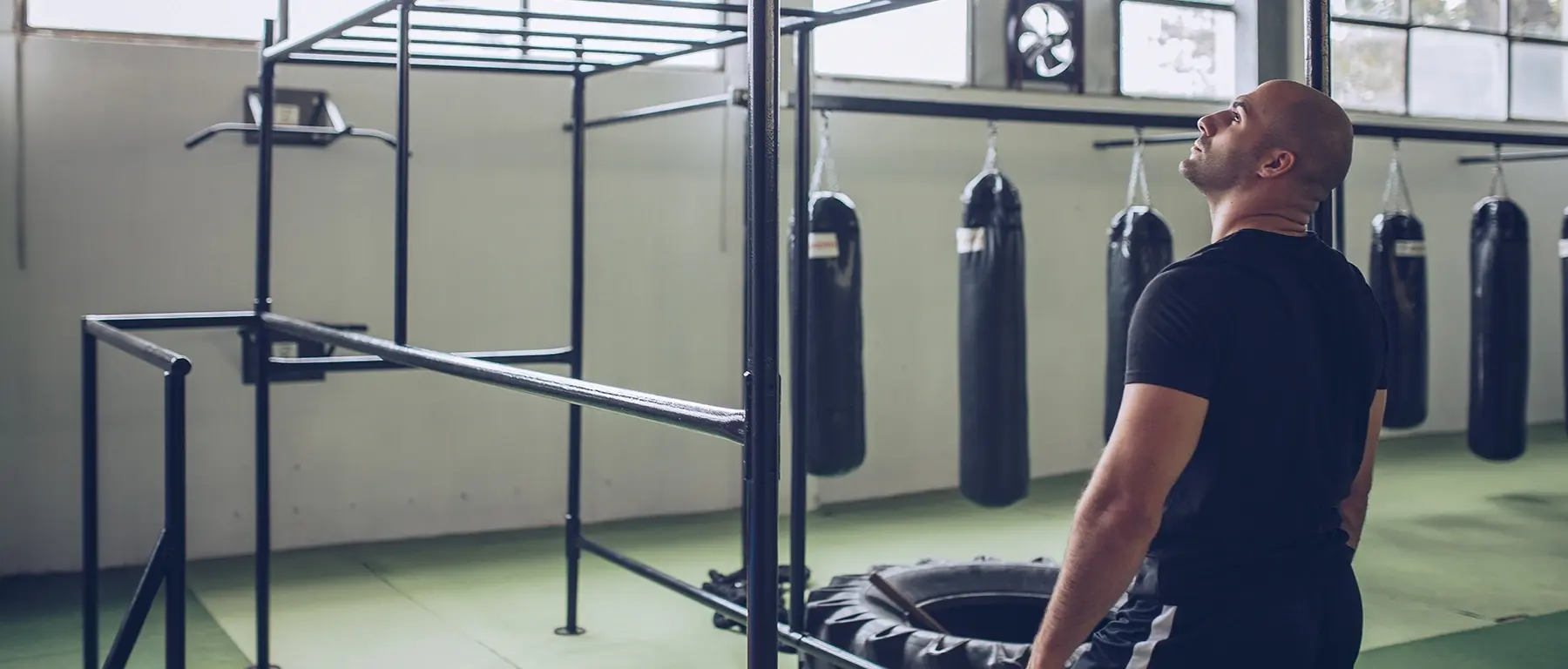 A man in a black t-shirt stands in a gym beside a squat rack and a large tyre, with a row of punching bags hanging behind him.