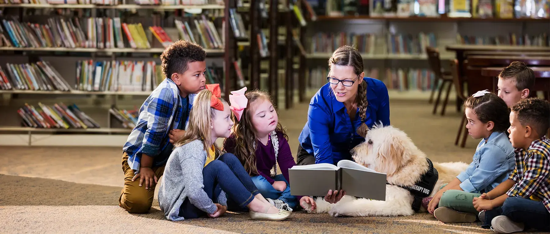 A woman reads aloud from a picture book to a group of young children sitting on a library floor, with a large white dog resting beside her.