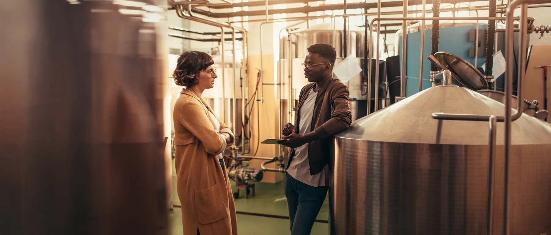 A woman and a man stand talking between large steel brewing tanks in a brewery, one holding a tablet.
