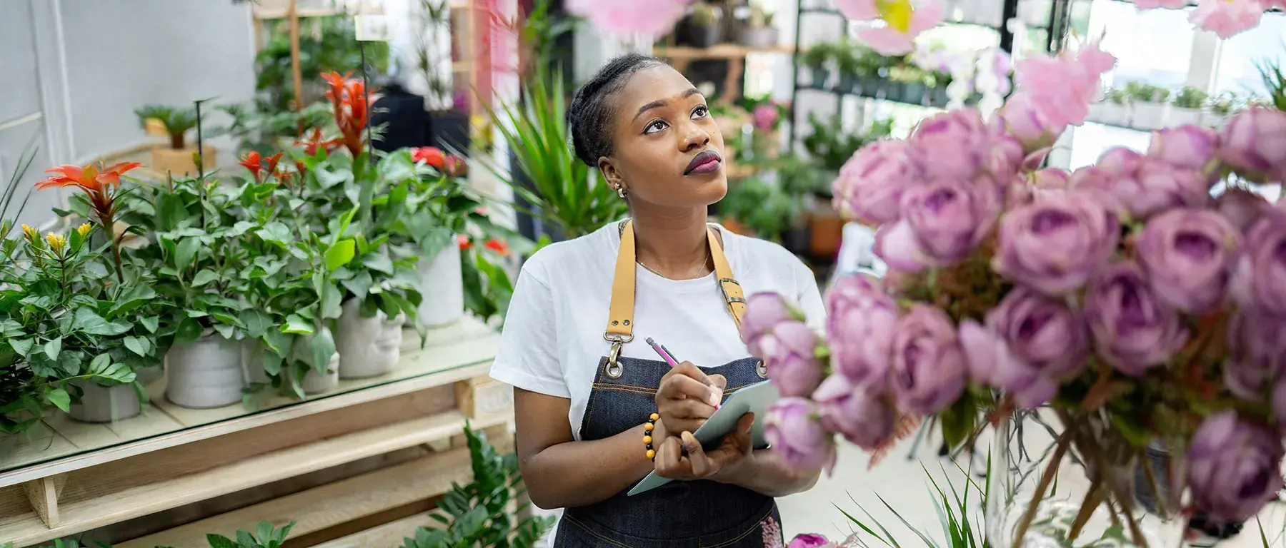 A woman in an apron writes on a clipboard among shelves of potted plants and pink roses in a flower shop.