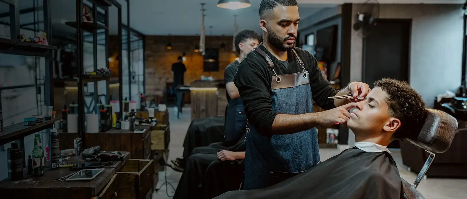 A barbershop owner trims a client's beard at his shop, with barber chairs and vintage decor in the background.