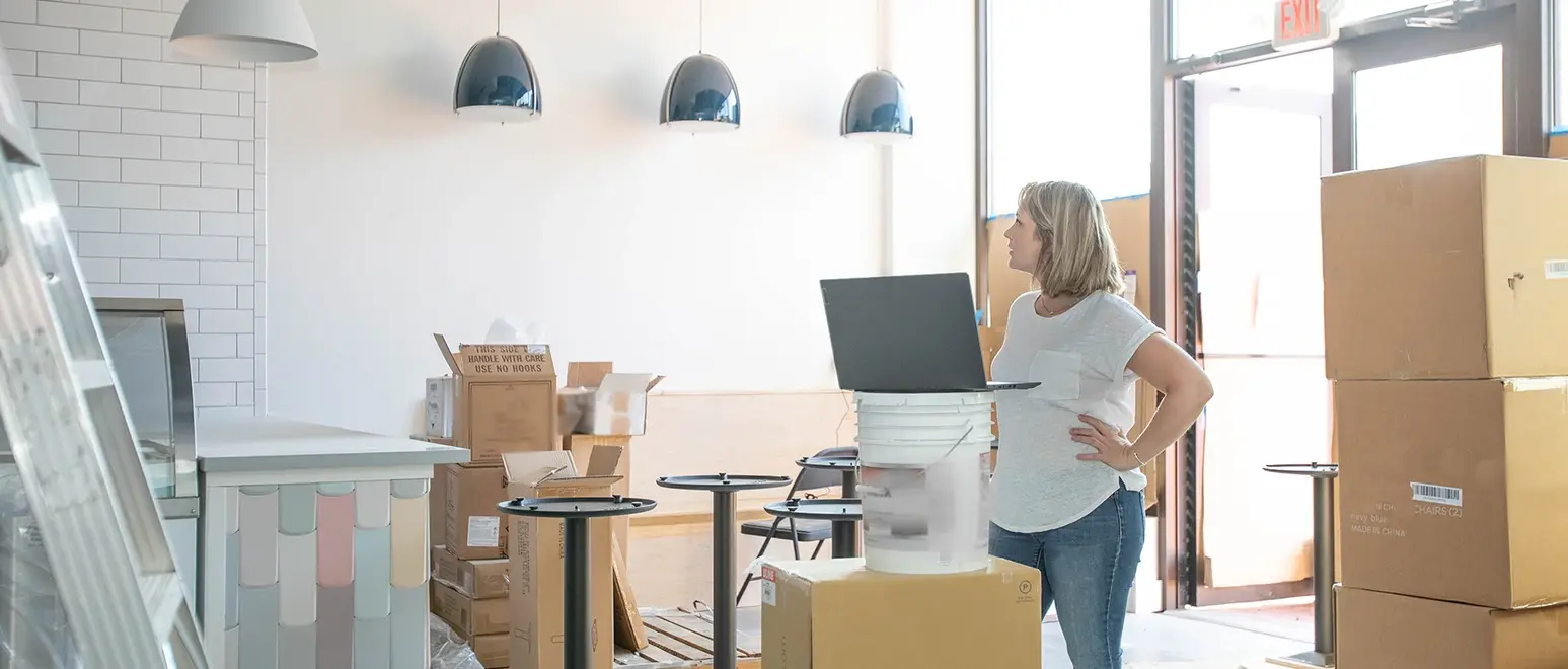 A small business owner reviews her laptop amid moving boxes in a bright storage or retail space.