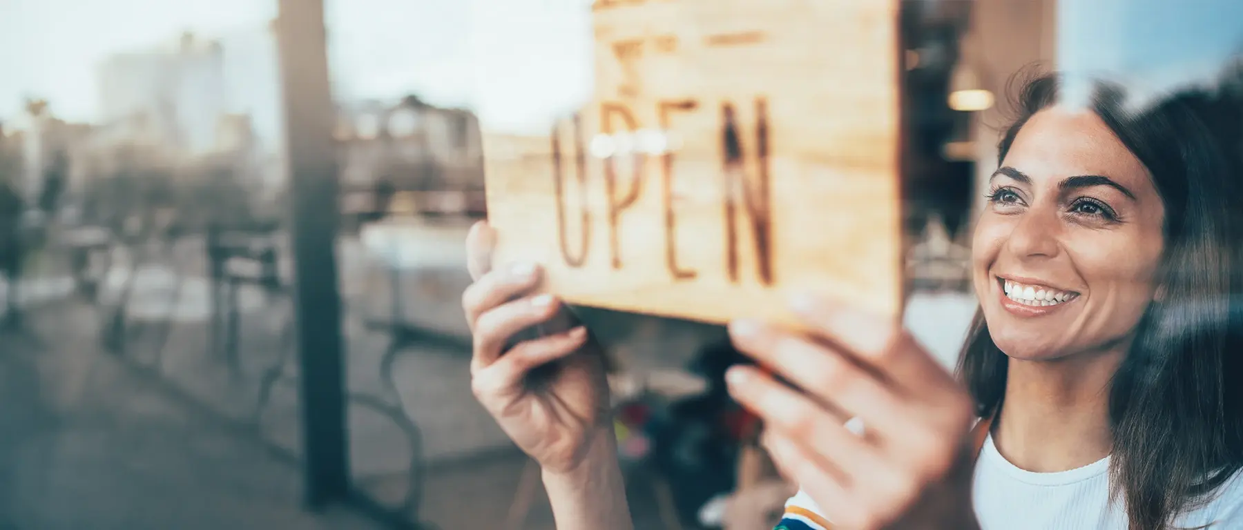 A smiling small business owner turns an OPEN sign in her shop front window, with a city street reflected in the glass.