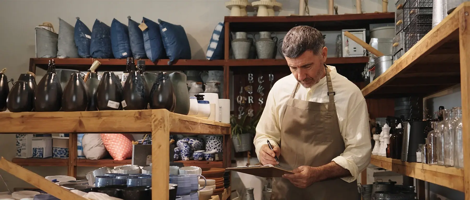 A man in an apron works at a wooden bench in a shop, with shelves of pottery, dark vases and folded textiles behind him.