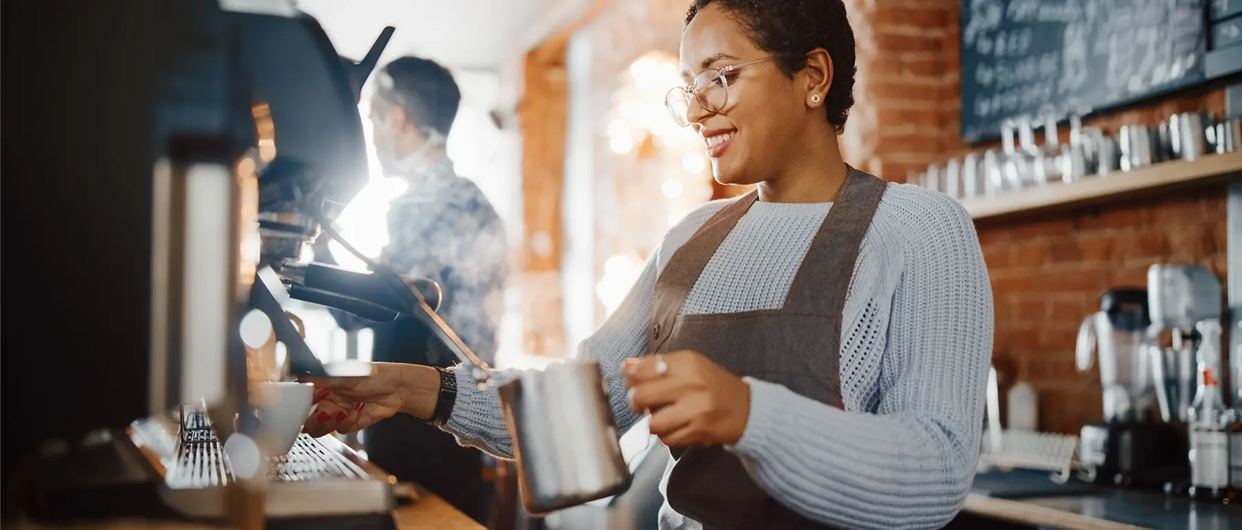 A smiling barista in an apron steams milk at an espresso machine in a café while a colleague works behind her.