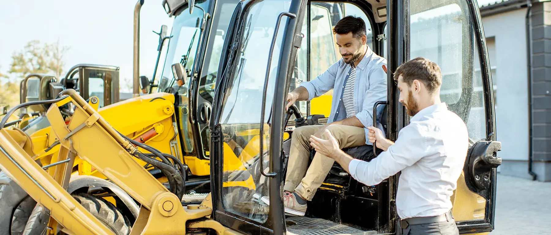 Two men in business clothes look over a document beside a row of yellow excavators, one of them seated in an open cab doorway.