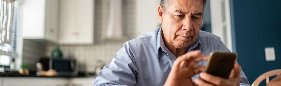An older man in a light blue shirt looks at his phone, with a kitchen visible behind him.