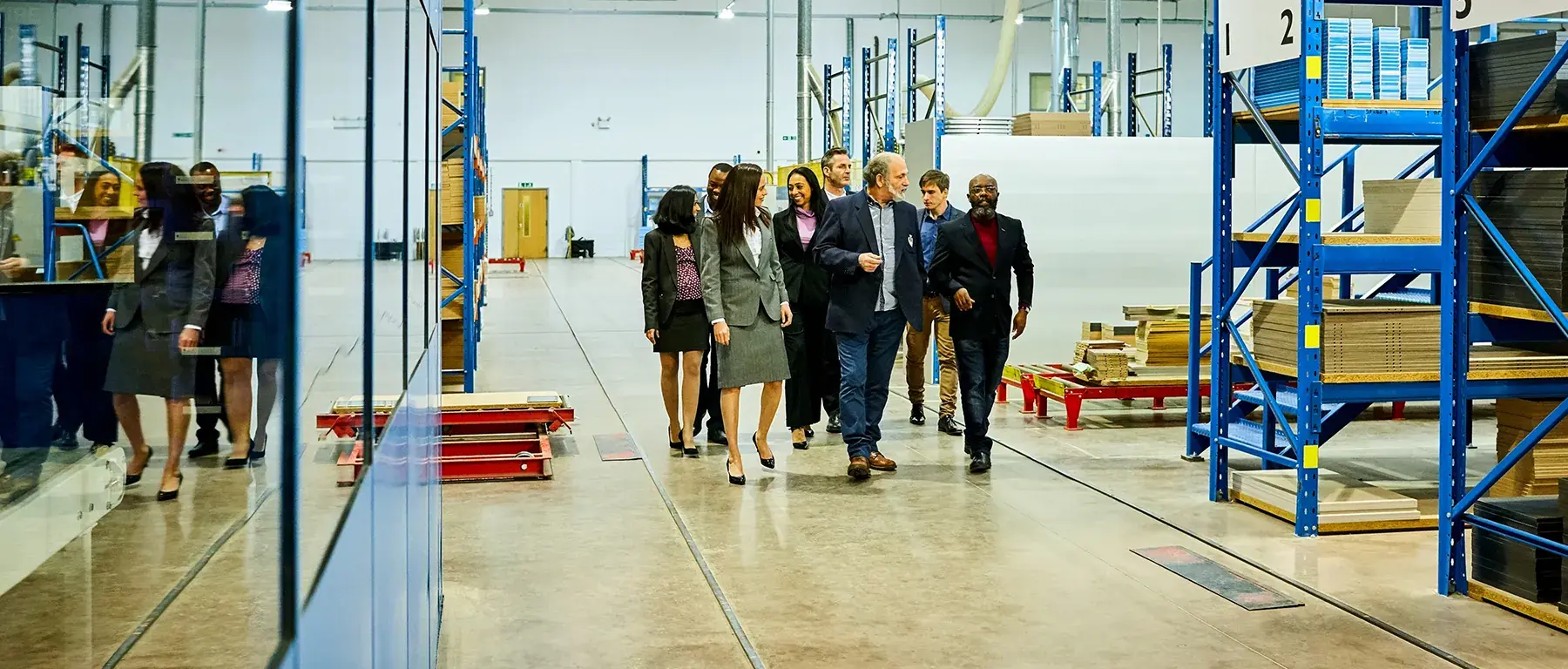 A group of business colleagues walks together down a warehouse aisle lined with blue steel racking and stacked pallets.