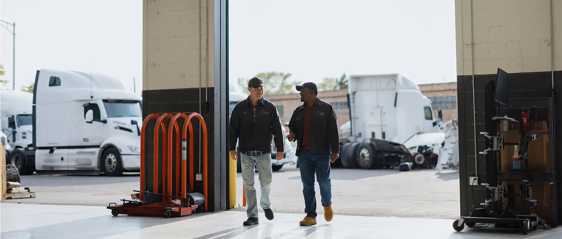 Two men in casual work attire walk past open bay doors at a trucking and logistics facility, semi-trucks parked outside.