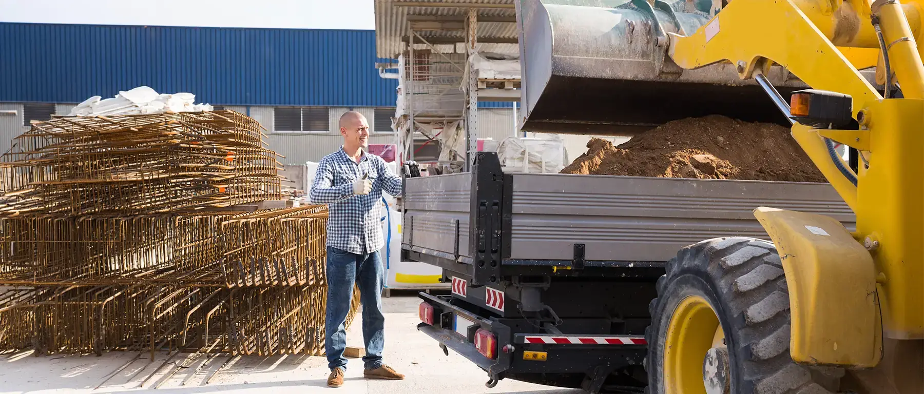 A man in a denim shirt stands beside rolled wire mesh and a loaded flatbed truck outside a construction supply warehouse.