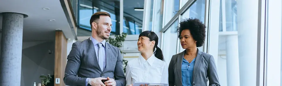 Three business professionals in suits walk and talk together in a bright, glass-walled office corridor.