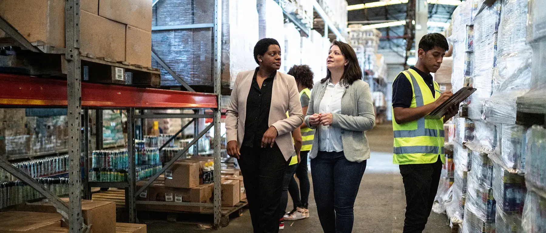 Three warehouse employees in high-visibility vests review inventory together among rows of packaged goods