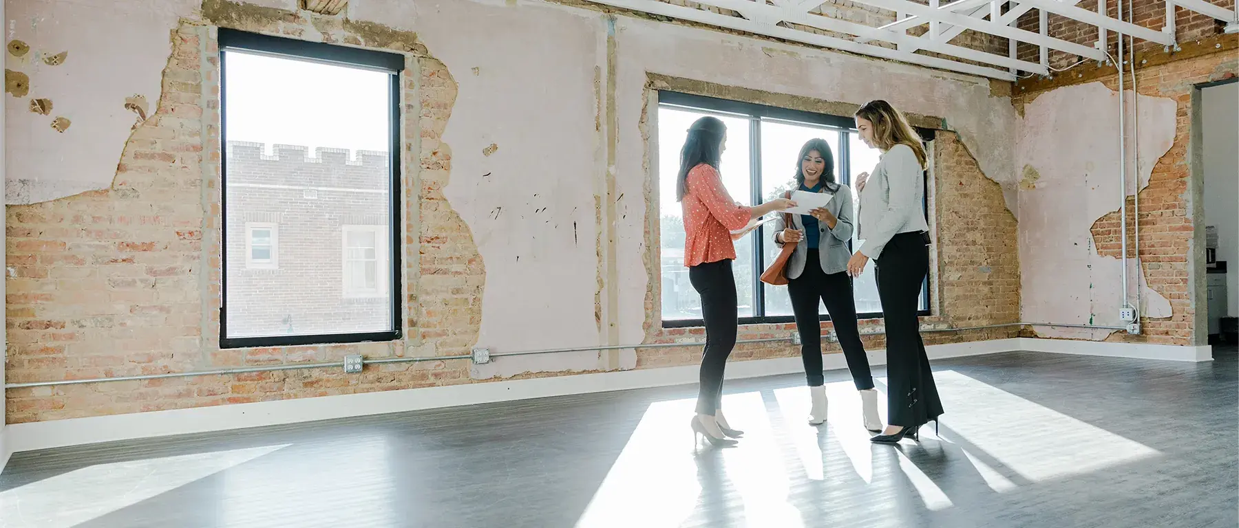 Three businesswomen discuss plans while standing in a bright, exposed-brick space under renovation