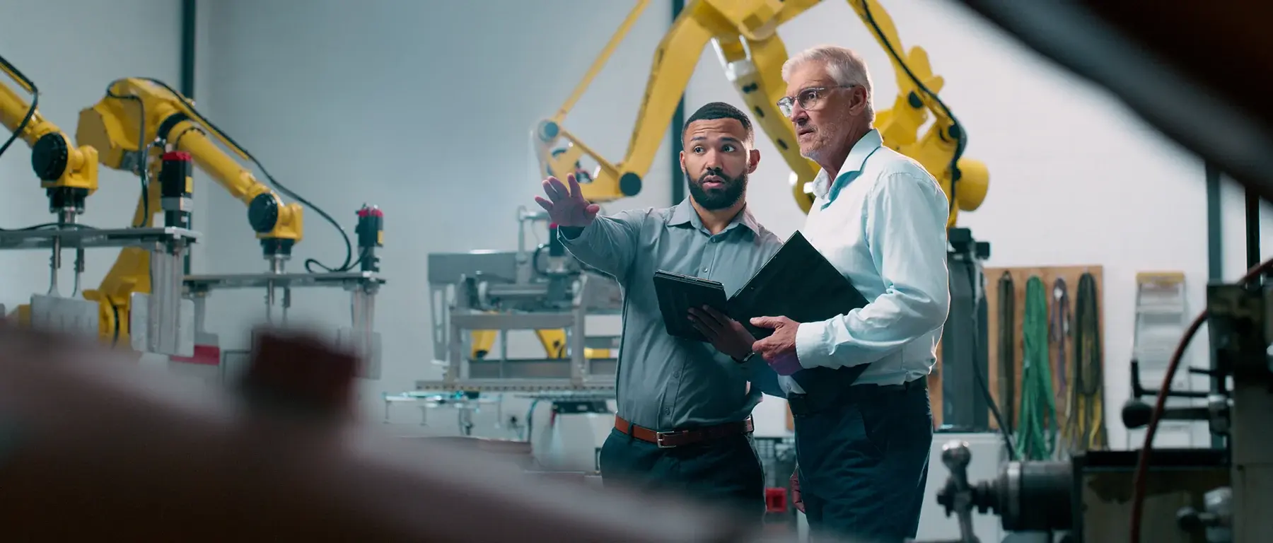Two businessmen in a manufacturing facility discussing plans near industrial robotic equipment