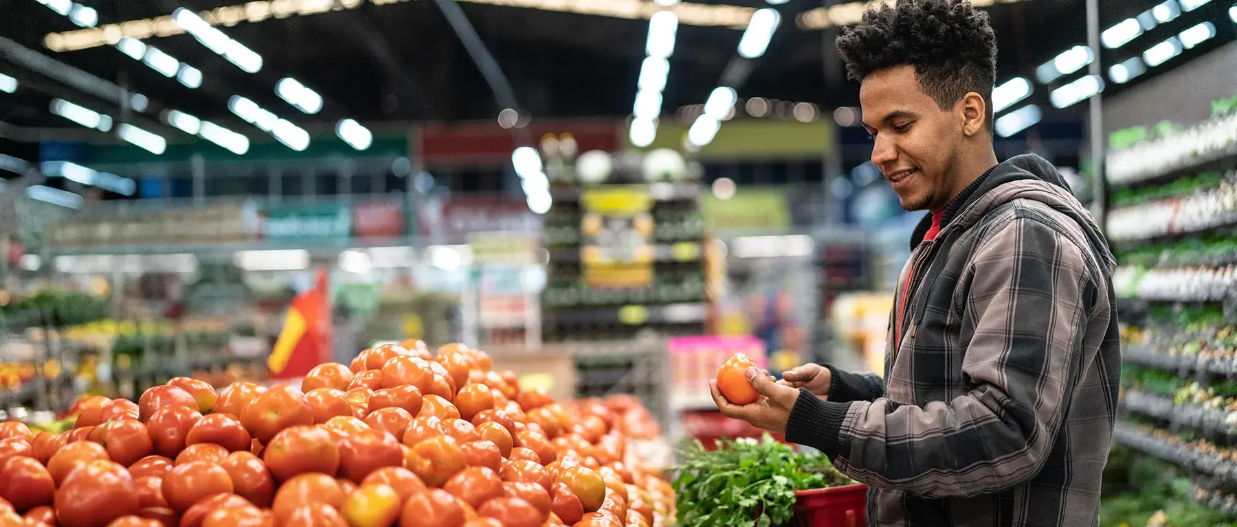 A man shopping in a grocery store selects a tomato from a large display of fresh produce.