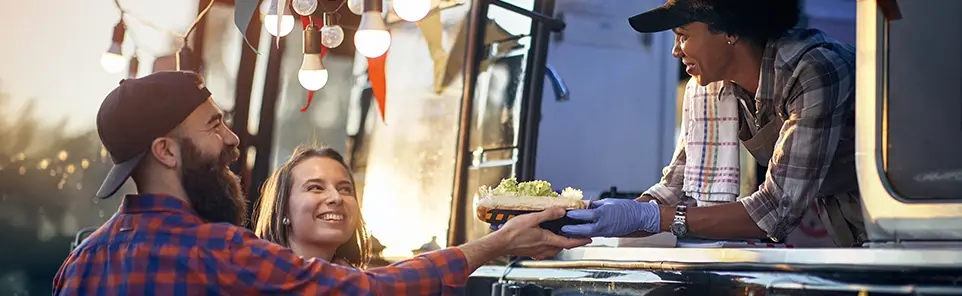 A smiling couple are handed a food order at a food truck window by a vendor in a cap.