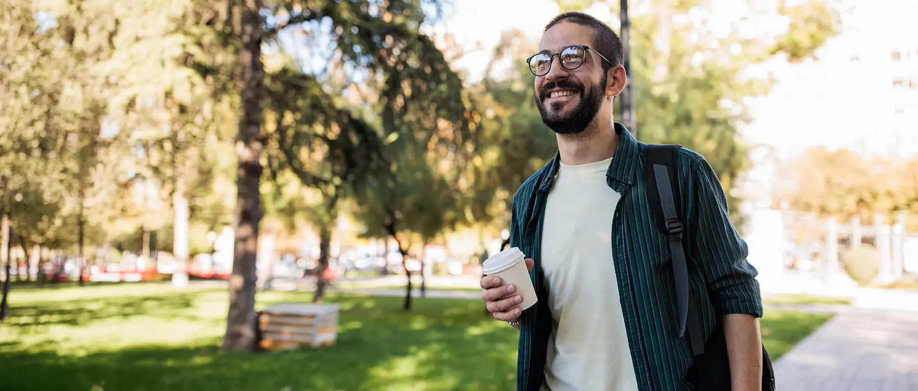 A smiling man with glasses and a backpack walks through a sunlit park holding a takeaway cup.