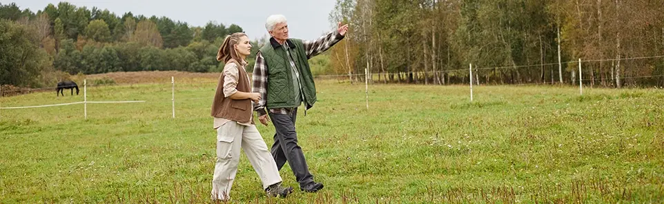 An older couple walk across a green paddock, one pointing into the distance past a fence line.