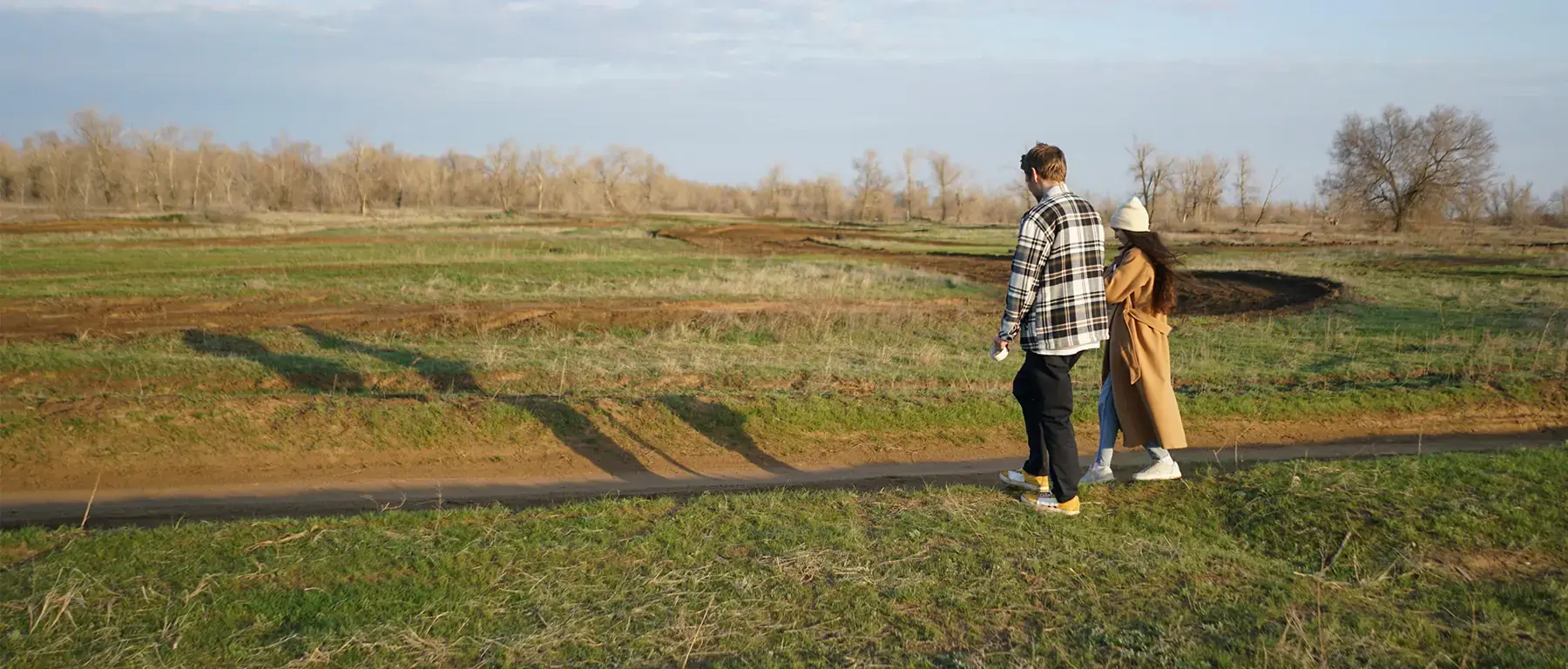 A couple walk along a track beside an open field of rough grassland under a pale sky.