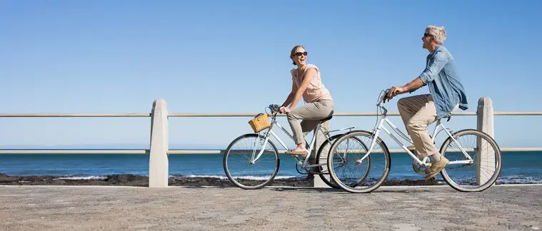 An older couple ride bicycles side by side along a seafront promenade on a clear day.