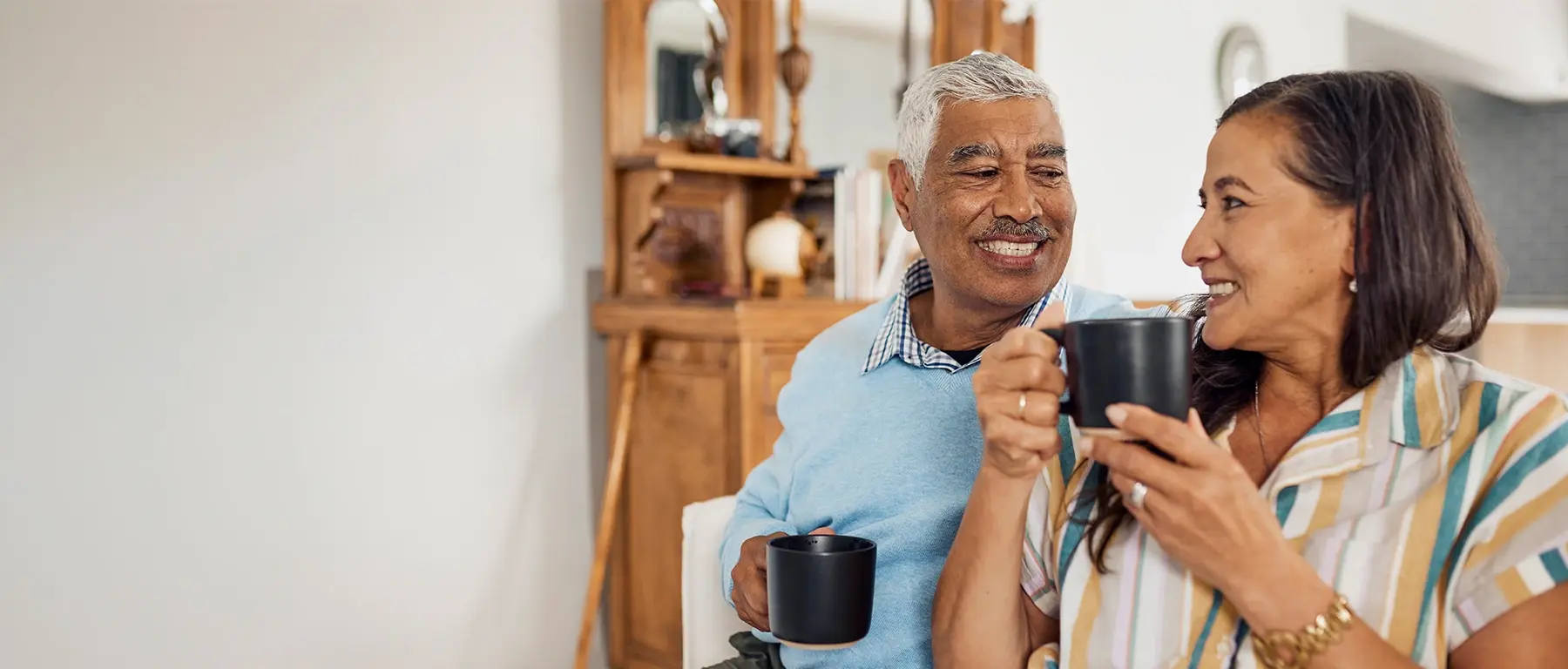 An older couple stand close together in a kitchen holding mugs of coffee, smiling at each other.