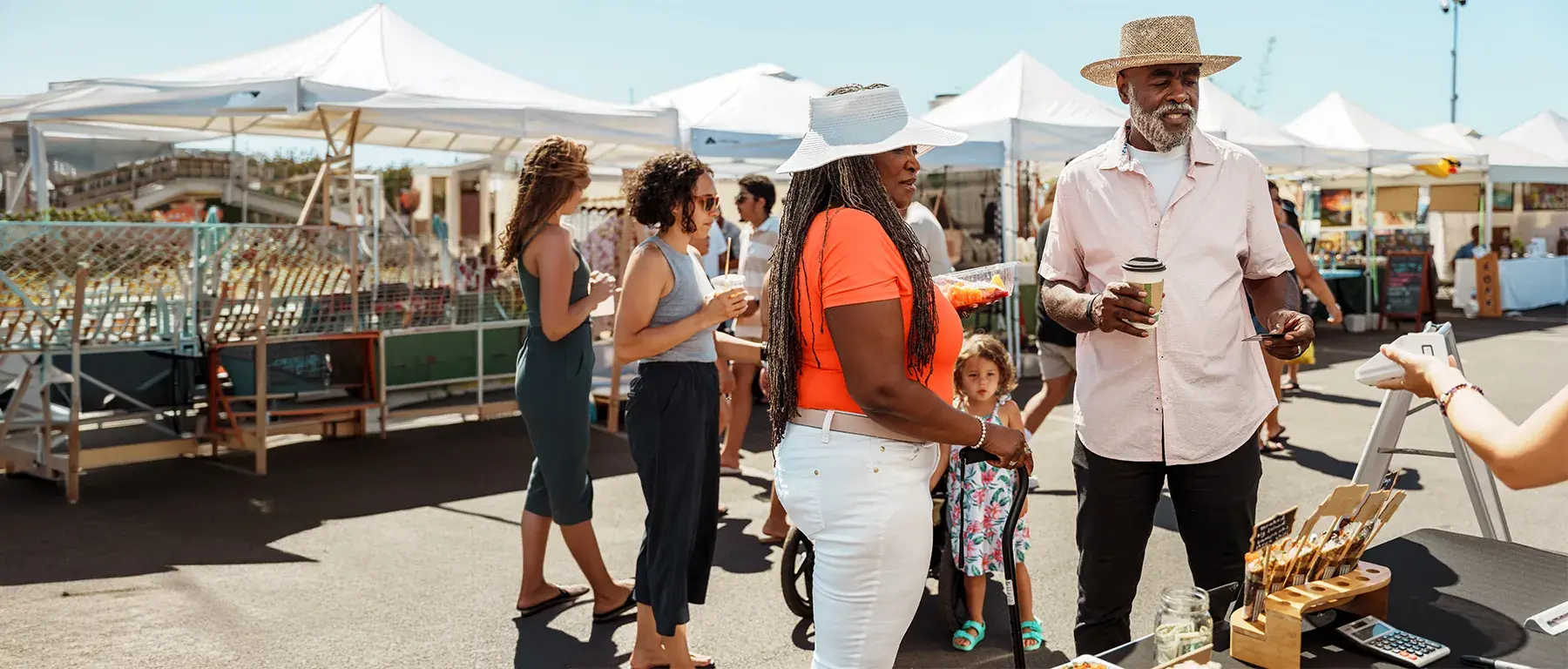 Shoppers browse stalls under white canopies at a sunny outdoor market, one holding an iced drink.
