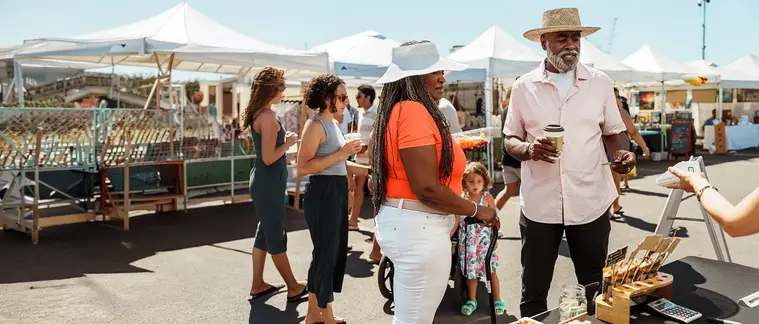Shoppers browse stalls under white canopies at a sunny outdoor market, one holding an iced drink.