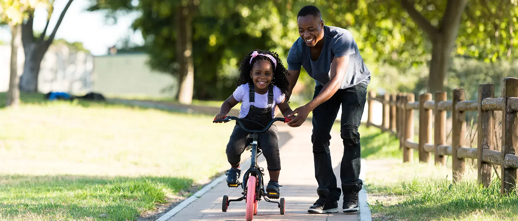 A man steadies a young girl learning to ride a bike along a sunlit path in a park.