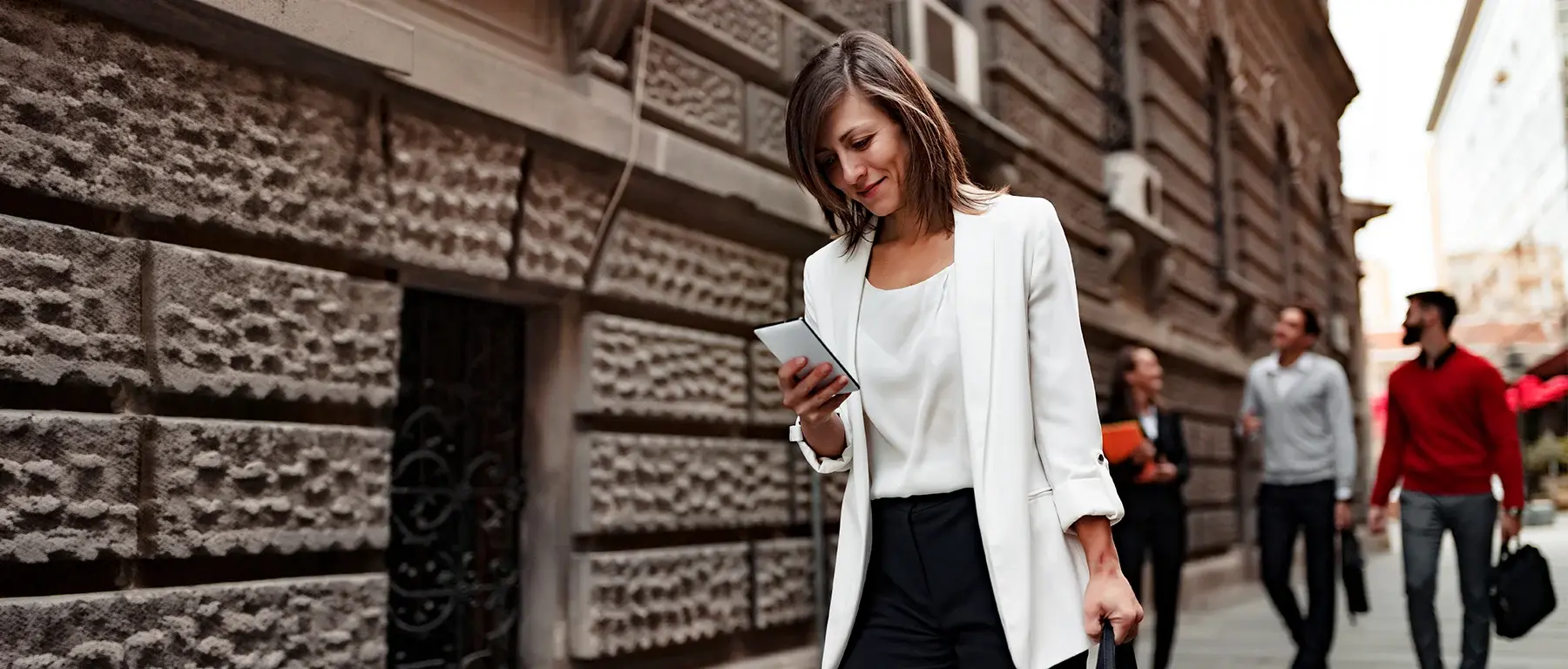 A woman in a white blazer checks her phone while walking down a city sidewalk, representing on-the-go account alert notifications.