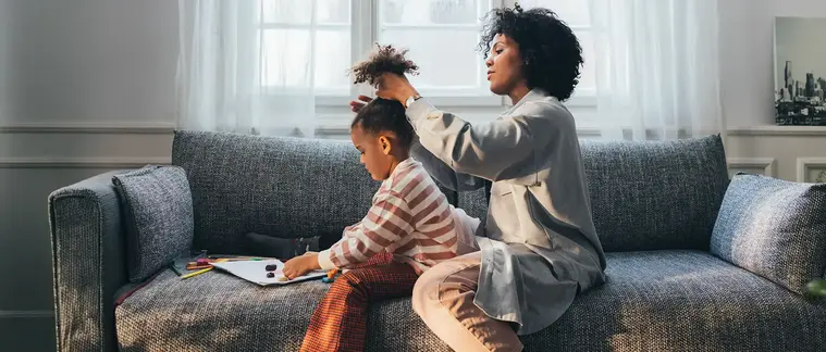 A woman sits on a grey sofa in a sunlit living room, tying back the hair of a young child who is drawing beside her