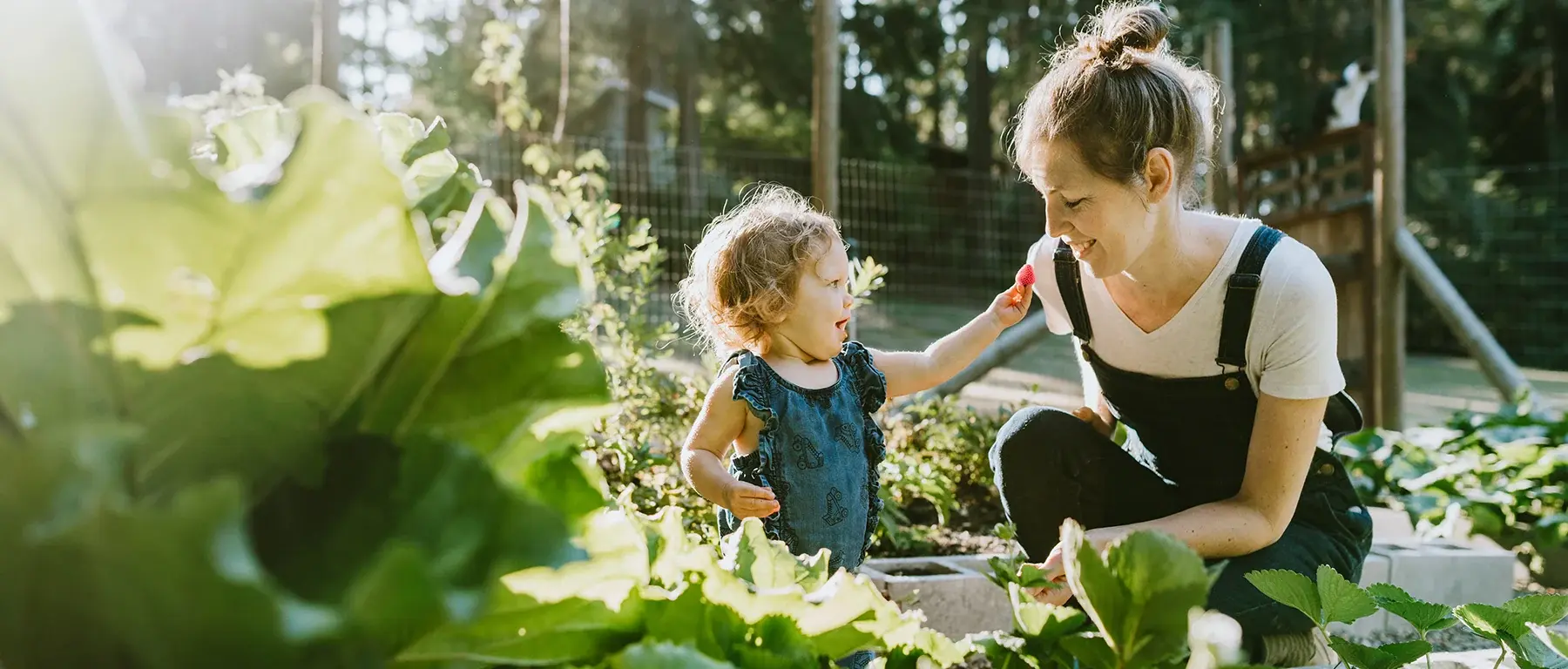 A woman in denim overalls crouches among leafy green plants in a sunlit garden, holding something out to a toddler beside her