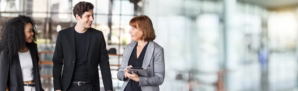 Three business professionals talk together in an office lobby, one holding a tablet.