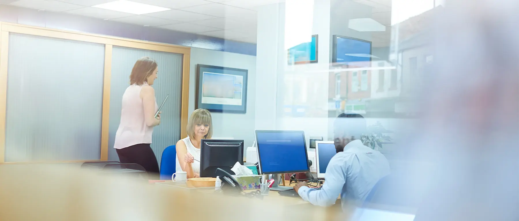 A manager reviews information on a screen with colleagues working at computer terminals inside a bright bank office.