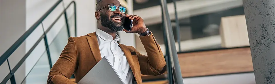 A smiling businessman in a blazer and sunglasses talks on his phone while holding a laptop on an outdoor staircase.